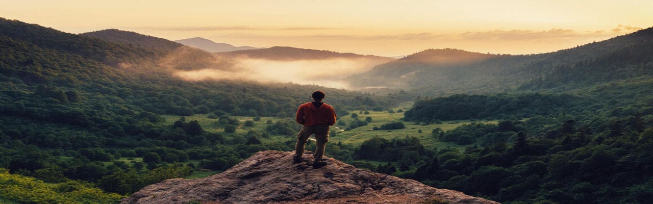 Young man standing on top of cliff in summer mountains at sunset and enjoying view of nature; Shutterstock ID 475421278; purchase_order: -; job: -; client: -; other: -