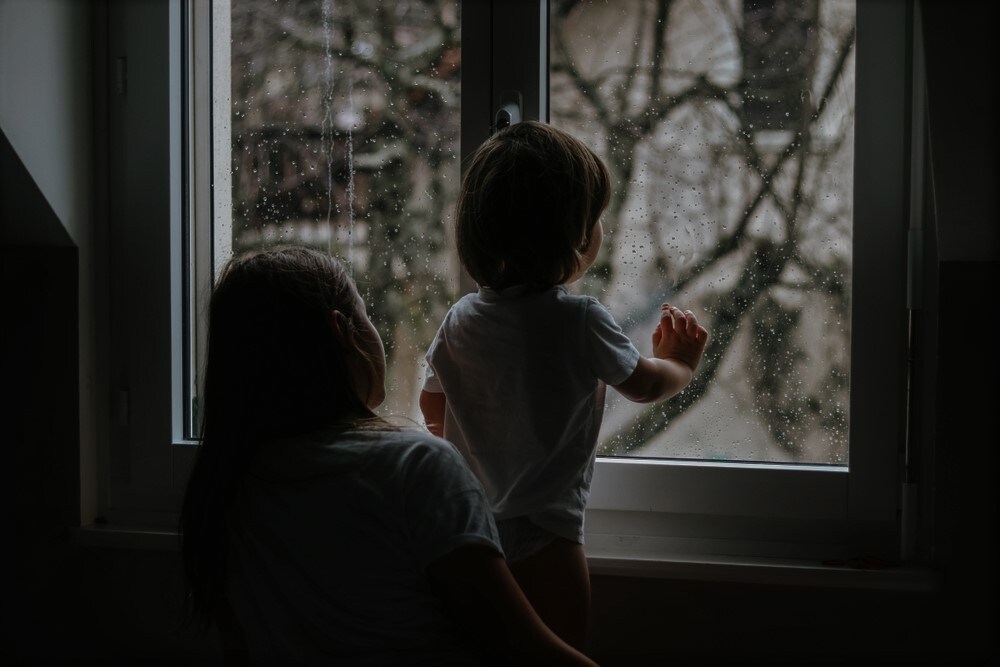 Rainy day. Little toddler boy with his sister looking outside through  wet window from his room missing mother.  Siblings love, care and support. Lifestyle. Quarantine and isolation period at home